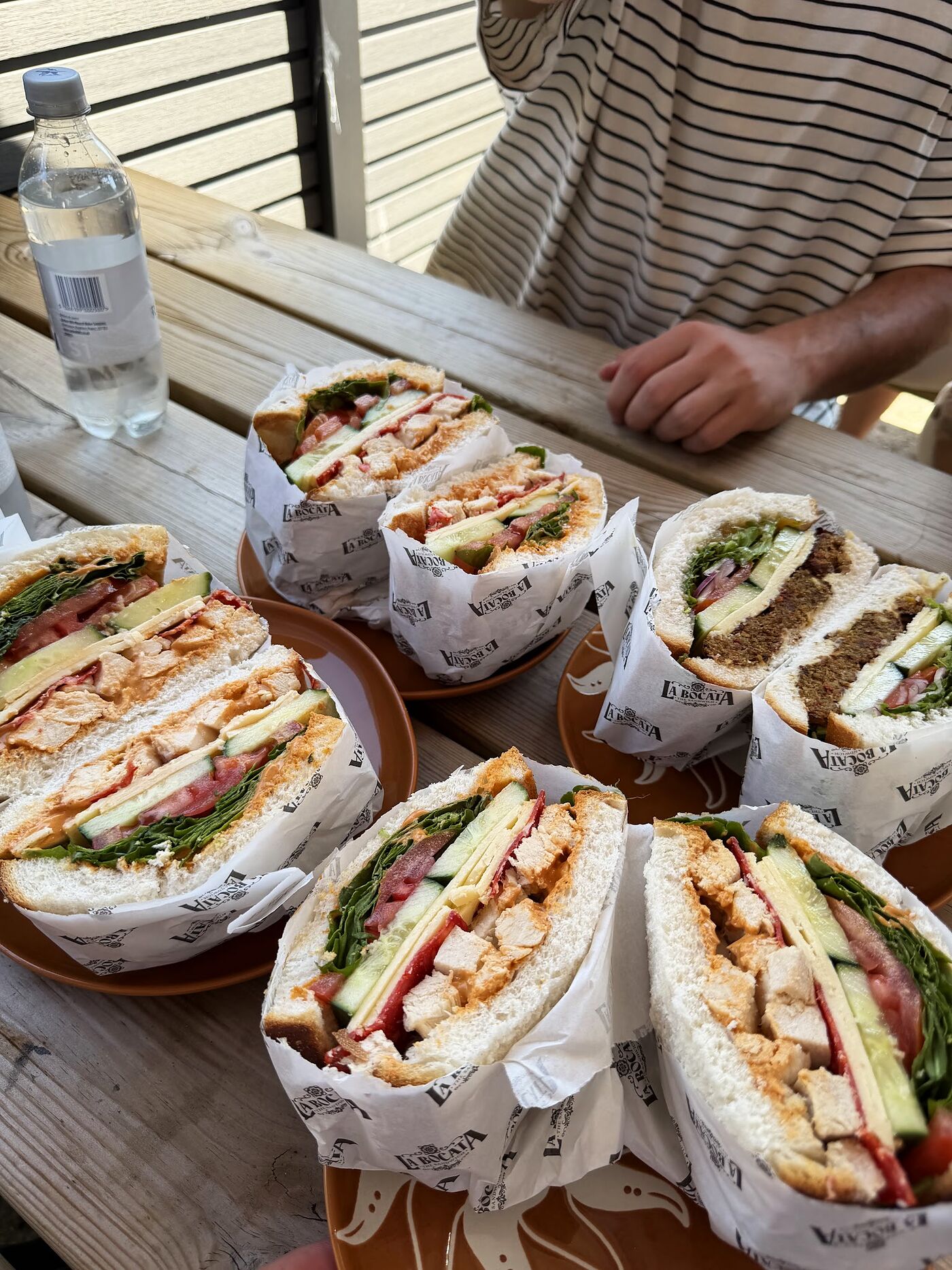 Half a dozen La Bocata sandwiches in branded wraps on terracotta plates, spread across an outdoor bench